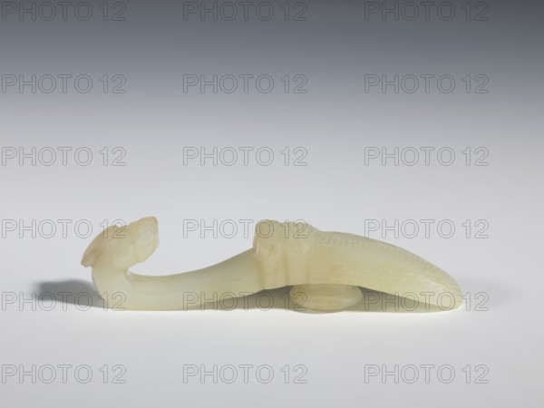 Belt clasp, Chinese, Qing dynasty 1644–1911, 18th–19th century. Milky-white translucent jade. Carved with cicadas and dragons; shaft in flat relief, head carved in full relief. Belt buckle; height 2.4 cm, width 10.7 cm, depth 2.1 cm., A pale, translucent carved object with a curved, elongated form rests on a flat surface, showing smooth and slightly glossy textures; one end features a rounded head-like shape with subtle carved facial details and a short raised element, while the opposite end tapers into a broader, textured section with shallow incised patterns and a ridged spine; a small rounded support sits beneath the midsection, casting a soft shadow, and the background transitions from light gray at the bottom to darker gray toward the top, with visible colors including off-white, cream, pale yellow, gray, and a hint of beige.