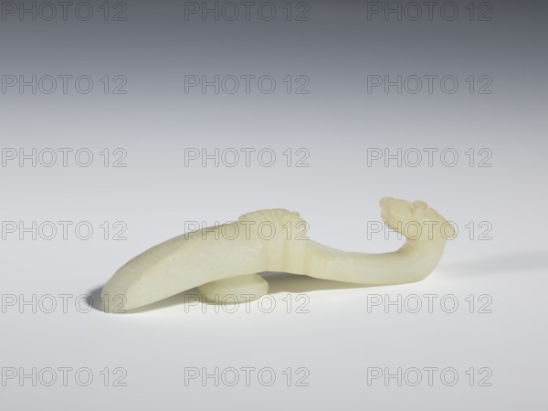 Belt clasp, Chinese, Qing dynasty 1644–1911, 18th–19th century. Milky-white translucent jade. Motifs: cicadas and dragons iconography. Shaft in flat relief; head carved in full relief. Belt buckle; height 2.4 cm, width 10.7 cm, depth 2.1 cm., A pale, light yellowish-white carved object resembling a curved form rests on a smooth, light gray surface against a gradient gray background, with a curved elongated body, a rounded underside support, subtle carved ridges along one edge, and a sculpted head-like end with rounded protrusions; shadows fall beneath and to the left of the object, and the overall scene shows shades of pale yellow, white, light gray, and darker gray in the background.