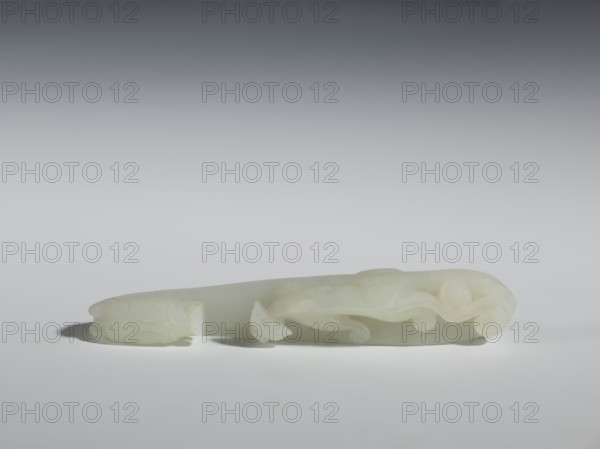 Belt clasp;, Milky-white, slightly translucent jadeite with slight colloid clusters. Carved and engraved; 18th–19th century Qing dynasty, China. Head shaped as a two-horned dragon’s head with an open mouth; handle features a lizard. Dragons, lizards iconography/zoology. Belt buckle. Height 2.1 cm; width 12.8 cm; depth 2.2 cm., A pale, translucent carved object rests horizontally on a smooth surface, showing a curved, elongated form with flowing, scroll-like raised details and rounded protrusions; the object appears to be a single pale greenish-white color with subtle variations, and it casts soft gray shadows on the light gray surface beneath, while the background transitions from light gray near the object to darker gray toward the top.