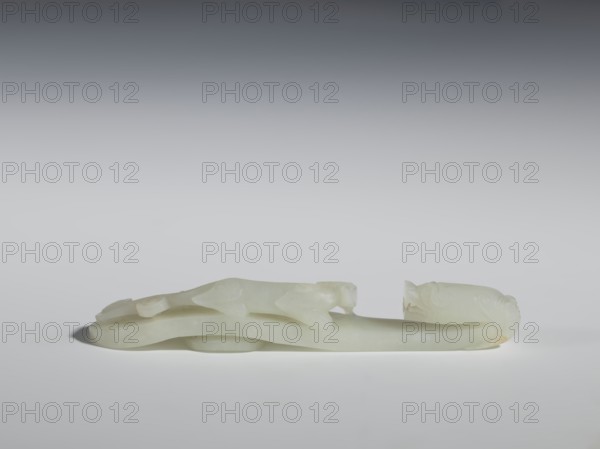 Belt clasp;, Chinese, Qing dynasty 1644–1911, 18th–19th century. Milky-white jadeite, slightly translucent with slight colloid clusters; carved and engraved. The head is in the shape of a two-horned dragon's head with an open mouth, and the handle features a lizard. Belt buckle; height 2.1 cm, width 12.8 cm, depth 2.2 cm., A pale, translucent carved object rests horizontally against a neutral background, showing a smooth, milky white surface with subtle areas of pale yellow and cream. The carving features flowing, elongated forms with rounded contours and gentle hollows, including a raised central section and curved end pieces that suggest stylized heads or decorative terminals. Soft shadows beneath the object create a sense of low elevation, and the overall palette includes shades of white, off-white, cream, and faint yellow against a gradient background ranging from light gray to darker gray at the top.