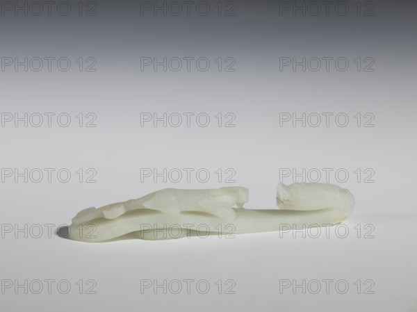 Belt clasp, Chinese, dragons iconography, lizards zool., Qing 1644–1911. The head is in the shape of a two-horned dragon's head with an open mouth, and the handle features a lizard. Milky-white jadeite, slightly translucent with slight colloid clusters. Carving, engraving, 18th–19th century, China, belt buckle. Height 2.1 cm, width 12.8 cm, depth 2.2 cm., A smooth, pale white carved object rests on a light surface against a neutral gradient background, showing an elongated, curved form with rounded ends and subtle raised details; the piece has soft translucent areas and slightly warmer off-white or cream patches, casting a faint shadow beneath it.