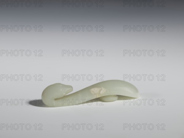 Belt clasp, Chinese, lizards zoological motif, Qing 1644–1911. Smooth, rounded handle. Milky-white jadeite, slightly translucent. Full-sculpture carved head, engraved. 17th–18th century, China. Belt buckle. Height 2.2 cm; width 10.5 cm; depth 2.1 cm., A smooth, curved object resembling an elongated animal form rests on a flat surface, showing a rounded head with subtle facial contours and a tapering body that loops gently; the object appears to be made of a polished, translucent material with a pale green color and a few slightly lighter patches, casting a soft shadow on the surface beneath, with the background transitioning from light gray near the surface to darker gray toward the top.