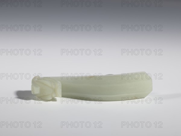 Belt clasp, stones; Chinese; dragons iconography; Qing 1644–1911, 18th–19th century; milky-white jade with colloid clusters, full-plastic carved head; belt buckle; height 2.1 cm, width 10.1 cm, depth 2.2 cm., A pale green, elongated carved object rests horizontally on a light surface against a gray gradient background, with one end featuring a carved rounded form resembling a stylized head and the opposite end faceted into an angled edge; the object casts a soft shadow and shows subtle variations of light green and off-white tones with faint darker spots.