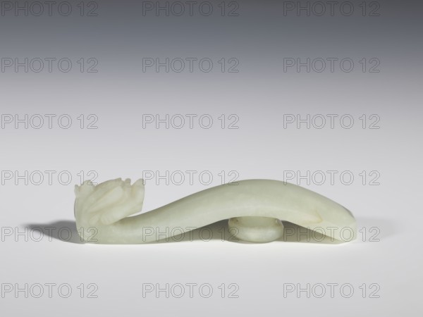 Belt clasp, stones, Chinese, dragons iconography, Qing style, Chinese art, Qing 1644–1911, milky-white jade with colloid clusters, full-plastic carved head, 18th–19th century, China, belt buckle, height 2.1 cm, width 10.1 cm, depth 2.2 cm, A pale greenish-white carved object rests on a smooth light-gray surface against a gradient gray background; the object has a curved, elongated body with a rounded arch supported by a small circular base and one end carved into a stylized floral or bud-like form with layered petal shapes, while the other end tapers to a flat, faceted tip, and a soft shadow falls beneath and to the left of the piece.