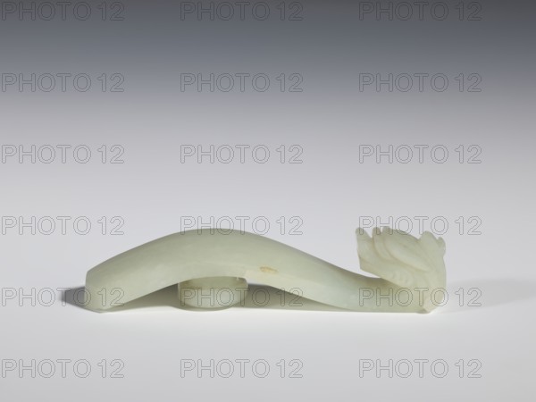 Belt clasp, stones, Milky-white jade with colloid clusters. Full-plastic carved head. Chinese, Qing dynasty 1644–1911, 18th–19th century. Dragons iconography. Belt buckle. Height 2.1 cm; width 10.1 cm; depth 2.2 cm., A pale green, smooth carved object with a curved elongated body resting on a round base and ending in a stylized carved head with layered, petal-like details, cast against a neutral gradient background in shades of gray and white, with soft shadows beneath the object and a small area of slightly darker discoloration on the side of the body.
