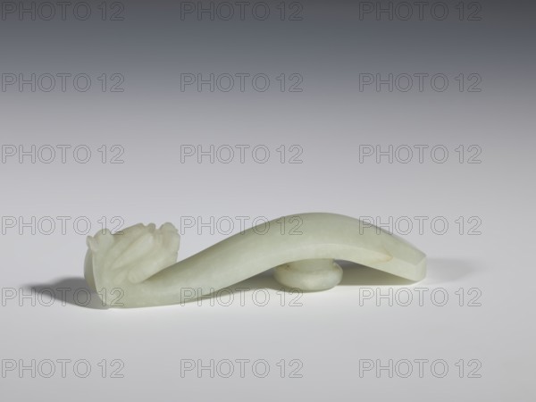 Belt clasp, stones; Chinese, Qing 1644–1911, 18th–19th century. Milky-white jade with colloid clusters; full-plastic carved head; dragons iconography. Belt buckle. Height 2.1 cm; width 10.1 cm; depth 2.2 cm., A pale green, curved carved object rests on a pale surface against a neutral gray gradient background; one end features a carved decorative cluster with rounded and petal-like forms and the other end tapers to a faceted edge, while a small rounded support sits beneath the arch casting soft shadows; visible colors include pale green, light gray, dark gray, and soft white.