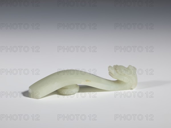 Belt clasp, stones; Chinese; dragons iconography; Qing style, Qing dynasty 1644–1911; milky-white jade with colloid clusters, full-plastic carved head; 18th–19th century, China; belt buckle; height 2.1 cm, width 10.1 cm, depth 2.2 cm., A smooth, pale green jade carving shaped like a curved object rests on a light surface, with a rounded support piece beneath its middle; one end tapers to a flattened tip while the other end features a carved floral or leafy motif with layered, rounded petals or leaves; soft shadows appear beneath and to the left of the object, and the background transitions from light gray near the surface to a darker gray toward the top.