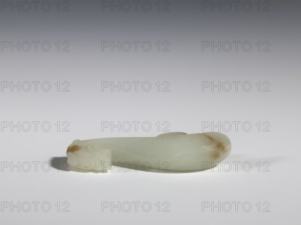 Belt clasp, Chinese, dragons iconography, Qing 1644–1911, milky-white jade with brown veins and clusters of colloids; full-sculpture carved head, engraved; 18th–19th century, China; belt buckle; height 1.9 cm, width 7.4 cm, depth 1.6 cm., A smooth, elongated object made of pale greenish-white stone with a few brownish discolorations rests horizontally on a flat, light-colored surface. The left end shows a carved, detailed section with layered, rounded forms and a small brown patch, while the right end tapers into a rounded, bulbous shape with a brownish area. Soft shadows fall beneath and to the left of the object, and the background transitions from light near the surface to darker gray toward the top.