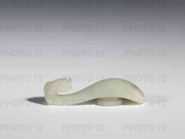 Belt clasp, Chinese, dragons iconography, Qing 1644–1911, milky-white jade with brown veins and clusters of colloids, full-sculpture carved head, engraved, 18th–19th century, China, belt buckle; height 1.9 cm, width 7.4 cm, depth 1.6 cm, A pale greenish-white carved object with a smooth, curved body and a carved end resembling a stylized head rests on a flat surface; the object casts a soft shadow and sits partly elevated on a small circular base. The background shows a smooth gradient from darker gray at the top to lighter gray near the surface, and the visible colors include pale green, white, gray, and subtle beige tones.
