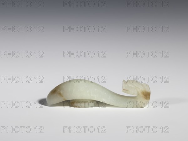 Belt clasp, Chinese, dragons iconography, Qing 1644–1911, milky-white jade with brown veins and clusters of colloids, full-sculpture carved head, engraved, 18th–19th century, China, belt buckle; H 1.9 cm, W 7.4 cm, D 1.6 cm., A pale greenish-white carved object with a smooth curved body and a carved end that resembles layered forms sits on a flat light surface; the carving shows areas of light brown veining and mottling, and a soft shadow falls beneath and to the left of the object against a background that transitions from light gray near the surface to darker gray toward the top.