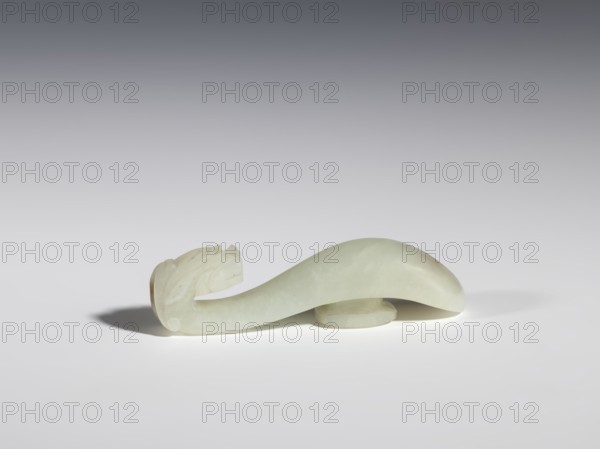 Belt clasp, Chinese, dragons iconography, Qing 1644–1911, milky-white jade with brown veins and clusters of colloids, full-sculpture carved head, engraved, 18th–19th century, China, belt buckle; H 1.9 cm, W 7.4 cm, D 1.6 cm., A smooth, curved jade object in pale green and off-white tones lies horizontally on a white surface, with a rounded elevated section beneath it and a carved end that resembles a stylized head, casting soft gray shadows on the surface under cool, neutral lighting and set against a gradient gray background.