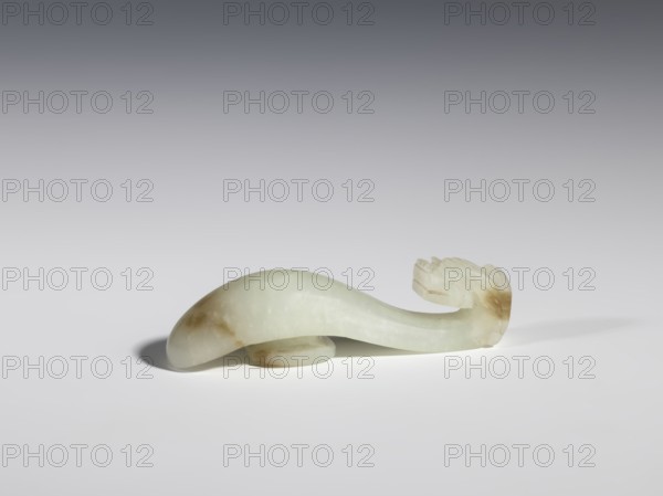 Belt clasp, Chinese, dragons iconography, Qing 1644–1911, milky-white jade with brown veins and clusters of colloids, full-sculpture carved head, engraved, 18th–19th century, China, belt buckle; height 1.9 cm, width 7.4 cm, depth 1.6 cm., An elongated, curved object carved from pale greenish-white stone with areas of light brown discoloration rests on a smooth white surface; one end is rounded and bulbous, the other tapers into a sculpted shape with layered ridges, and a soft shadow appears beneath and to the left of the object against a background that transitions from light gray near the surface to darker gray toward the top.