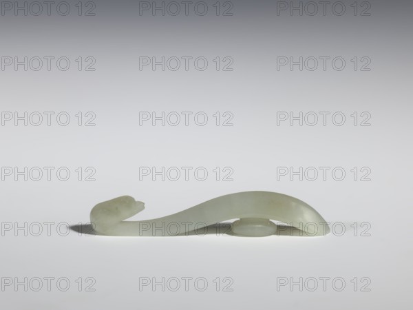 Belt clasp, Chinese, birds, Qing 1644–1911, milky-white jadeite with slight green tint, full-sculpture carved head, handle with an edge on the spine and a pointed tip, 17th–18th century, China, belt buckle; h. 1.3 cm, w. 7.3 cm, d. 0.96 cm., A smooth, curved object resembling a stylized snake or serpentine form rests on a flat surface, carved from a pale green, almost translucent stone with subtle variations in hue; one end rises slightly with a rounded head-like shape and a small open mouth, while the body arches gracefully and is supported near the center by a low, disc-like element, casting soft gray shadows on the white-gray background.