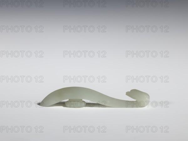 Belt clasp, Chinese, Chinese art, birds, Qing 1644–1911, handle with an edge on the spine and a pointed tip. Milky-white jadeite with clusters of colloids and a slight green tint. Full-sculpture carved head. 17th–18th century, China, belt buckle, height 1.3 cm, width 7.3 cm, depth 0.96 cm., A pale green, smooth carved object with a curved, sinuous form resembling a stylized animal, positioned horizontally on a flat white surface; the object casts a soft shadow beneath and has a rounded head at one end with subtle carved features, a raised arch in the middle supported by a small oval base, and a tapered tail at the other end; the background transitions from light gray near the surface to a darker gray toward the top.