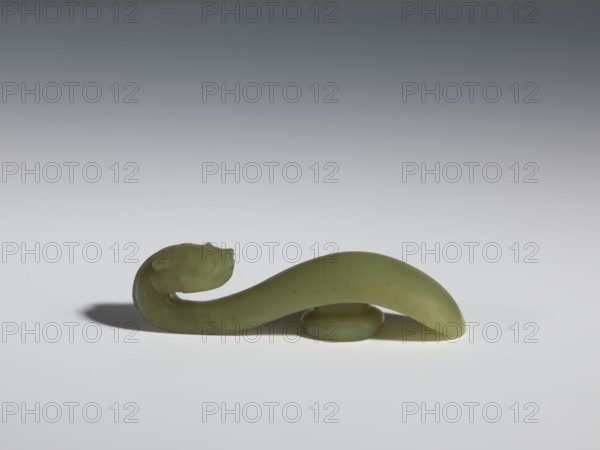 Belt clasp, Chinese, Qing 1644–1911, 17th–18th century. Jadeite with a green shade and visible clusters of colloids. Head carved in full relief, engraved; dragons iconography. Belt buckle. Height 2.2 cm, width 8.5 cm, depth 2.25 cm., A smooth, curved green object with a glossy surface shaped like a sinuous form rests on a flat pale surface, its thinner end curling upward into a rounded head-like shape and the broader end arching over a small rounded support; the object casts a soft shadow, and the visible colors include shades of green, pale gray, and darker gray in the background.