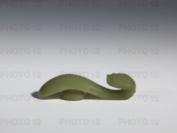 Belt clasp, Chinese, dragons iconography, Qing style, Qing 1644–1911. Jadeite with a green shade with visible clusters of colloids. Head carved in full plastic, engraved, 17th–18th century, China. Belt buckle; H 2.2 cm, W 8.5 cm, D 2.25 cm., A smooth, curved object in a pale green color rests on a white surface with a soft gray gradient background, showing a rounded, elongated form that arches over a small circular base and tapers into a sculpted end with subtle carved details; the scene includes soft shadows beneath the object and variations of green and gray tones.