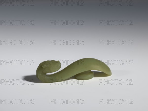 Belt clasp, Chinese; dragons iconography; Qing 1644–1911, 17th–18th century; jadeite with a green shade with visible clusters of colloids; head carved in full plastic, engraved; belt buckle; height 2.2 cm, width 8.5 cm, depth 2.25 cm., A smooth, curved green object resembling a sculpted loop with a rounded end sits on a light surface, casting a soft shadow; the piece has a slightly darker green hue in places and a matte finish, and the background transitions from light gray near the surface to darker gray toward the top.