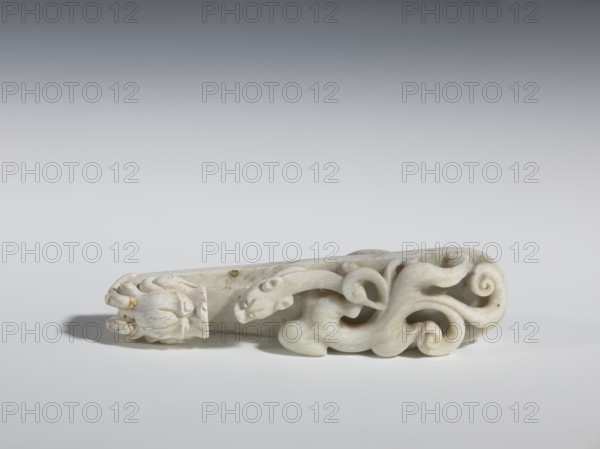 Belt clasp. Handle: stylized lizard-dragon qilong. Milky-white jadeite. Full-plastic carved head, full-plastic openwork handle. 18th–19th century, China. Belt buckle. Height 2.8 cm, width 12.0 cm, depth 2.9 cm. Subjects/themes: Chinese art; Qing 1644–1911; men's clothing, ornaments; lizards, dragons., A pale stone carving lies horizontally on a light surface, showing a sculpted form with flowing, curled shapes resembling stylized waves or tendrils on one end and a rounded, bud-like element on the other; the stone is mostly off-white with subtle gray veining and small darker specks, and there are faint shadows beneath the object with a gentle gradient background shifting from light gray at the bottom to darker gray toward the top.