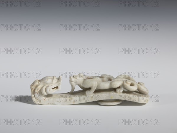 Belt clasp. Handle: stylized lizard-dragon qilong. Milky-white jadeite. Full-plastic carved head, full-plastic openwork handle. 18th–19th century, China. Belt buckle; height 2.8 cm, width 12.0 cm, depth 2.9 cm. Cultural context: Chinese, Qing 1644–1911. Subject/ornamentation: lizards, dragons; men's clothing/ornaments., A carved pale stone object with a smooth, slightly yellowed surface shaped into a long, low form; one end features a stylized animal head with an open mouth and curved ear-like elements, and the body rises into a sinuous, looping creature with rounded limbs and a curled tail resting along the top; the base is flat with a small circular support underneath, and the piece shows subtle speckles and veins in tones of white, cream, beige, and faint yellow against a neutral gray background.