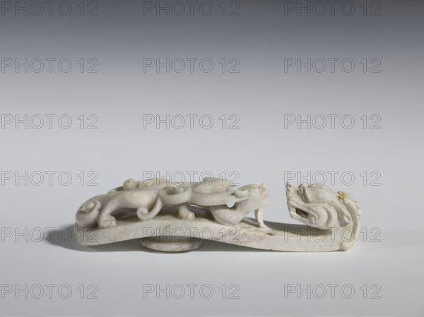 Belt clasp. Handle: stylized lizard-dragon qilong. Milky-white jadeite. Full-plastic carved head, full-plastic openwork handle. 18th–19th century, China. Belt buckle. Height 2.8 cm; width 12.0 cm; depth 2.9 cm. Categories/subjects: Chinese art; Qing 1644–1911; clothing; belts; men's clothing; costumes; ornaments; men's ornaments; lizards; buckles; dragons iconography., A carved object made of pale white stone with subtle beige and gray veining rests on a neutral gray surface, showing a sinuous form with curled and rounded decorative elements and a raised curved base; the piece features sculpted scroll-like shapes and flowing contours, with smooth and slightly weathered texture and small patches of darker beige discoloration, set against a gradient gray background that shifts from lighter at the bottom to darker at the top.