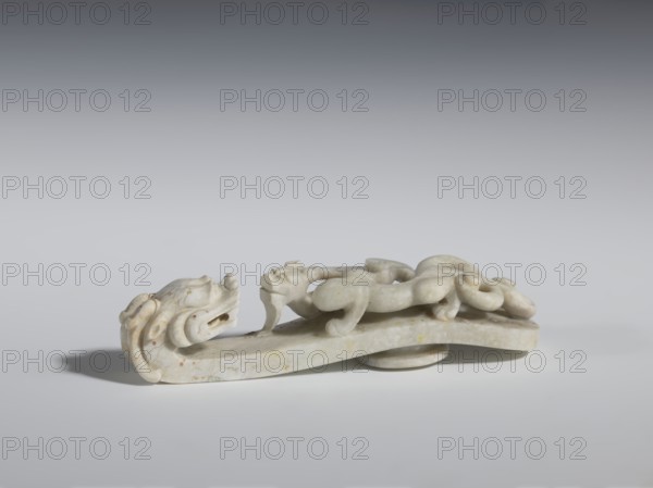 Belt clasp. Handle: stylized lizard-dragon qilong. Milky-white jadeite. Full-plastic carved head, full-plastic openwork handle. 18th–19th century, China. Belt buckle; height 2.8 cm, width 12.0 cm, depth 2.9 cm. Materials/subjects: Chinese, Qing 1644–1911, men's clothing, belts, ornaments; motifs: lizards/dragons., A pale stone carving shaped like a horizontal elongated base supports a sculpted creature with a curled tail and rounded limbs; the creature and base are the same off-white to light beige color with subtle yellowish and brown speckles and veins, and the surface has smooth, slightly weathered contours. The piece casts a soft shadow on a neutral gray surface and is set against a gradient background ranging from light gray near the object to darker gray toward the top. The carving shows flowing, rounded forms and gentle grooves that suggest stylized features, with the overall palette limited to shades of off-white, beige, yellowish spots, brownish flecks, and various grays in th...
