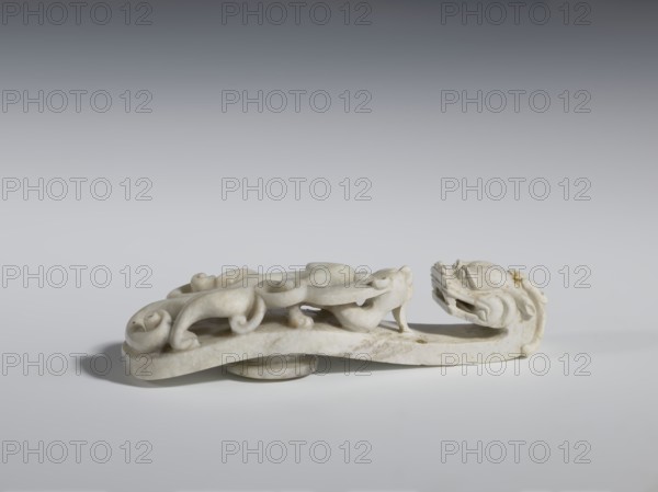 Belt clasp. Handle: stylized lizard-dragon qilong. Milky-white jadeite. Full-plastic carved head; full-plastic openwork handle. 18th–19th century, China. Belt buckle; height 2.8 cm, width 12.0 cm, depth 2.9 cm. Categories: Chinese art; Qing 1644–1911; men's clothing; costumes; ornaments; men's ornaments; lizards; dragons., Purchase, A low, elongated carved object with a stylized animal head at one end and a sinuous, scrolling body along its length, rendered in pale off-white stone with subtle areas of darker discoloration and small brownish spots; the piece sits on a flat surface casting a soft gray shadow and is photographed against a smooth gradient background ranging from light gray at the bottom to darker gray toward the top.