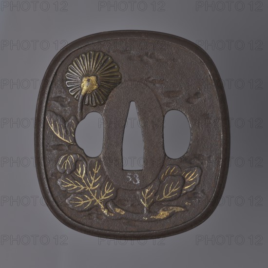 Crossguard tsuba, Tôryûsai school; 19th century Edo period, 1603–1868, Japan. Tsuba in the shape of a rectangle with rounded sides and corners nadegaku gata, slightly raised with a rounded profiled edge. Surface decorated in low and high relief: obverse shows a large butterfly in flight seen sideways, with a nakago-ana through its wings, an oval seppa-dai, and side hitsu-ana; details engraved and highlighted with gilded nunome inlay. Reverse depicts a chrysanthemum sprig with a flower visible from the back, rendered in brass high-relief inlay takazôgan and gold nunome inlay. Unsigned. Materials: metal with brass and gold inlay. Type: tsuba sword crossguard. Dimensions: height 8 cm, width 7.5 cm, depth 0.6 cm., A dark brown rounded square metal object with a central vertical oval opening flanked by two rounded openings, decorated with a raised flower motif at the upper left and a group of leafy plant motifs along the lower edge, with several gilded highlights on parts of the flower and ...