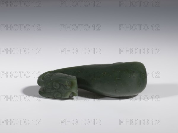 Belt clasp, Chinese, Chinese art, dragons iconography, Qing 1644–1911, clothing, belts, men's clothing, costumes, ornaments, men's ornaments, buckles, spinach jade, uniformly colored, full-plastic carved head, engraving, bas-relief, 18th–19th century, China, belt buckle; height 3.5 cm, width 9.5 cm, depth 3.1 cm, A smooth, dark green stone object rests on a light surface with a soft shadow beneath it; one end of the object features carved decorative swirls and rounded shapes, while the rest tapers into a polished, irregularly rounded form with small darker flecks; the background transitions from pale gray near the surface to a darker gray toward the top, and visible colors include shades of dark green, light gray, and darker gray.