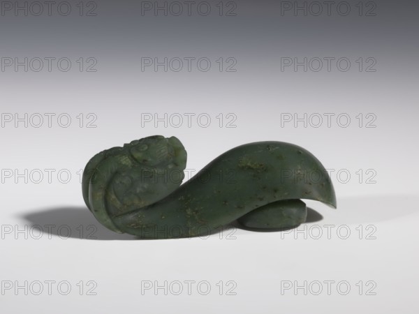 Belt clasp, Chinese, dragons iconography, Qing 1644–1911, men's clothing, belts, costumes, ornaments, men's ornaments, buckles, spinach jade, uniformly colored, full-plastic carved head, engraving, bas-relief, 18th–19th century, China, belt buckle; height 3.5 cm, width 9.5 cm, depth 3.1 cm, A small carved object made of dark green stone with lighter green and brown speckles rests on a smooth light surface, casting a soft shadow; the carving has a curved, flowing shape with one end forming a detailed, rounded figure with carved ridges and swirl-like patterns and the other end tapering to a smooth, hooked curve.
