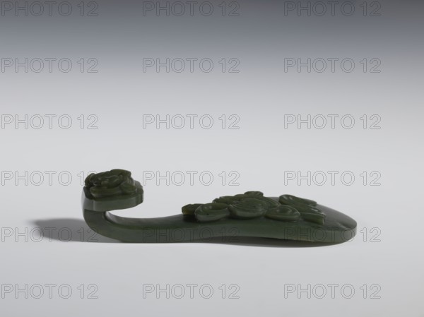 Belt clasp, Chinese, lizards zool., ruyi auspicious scepter iconographic, Immortals iconographic, Taoism, Qing 1644–1911. Scepter head with a bas-relief stylized lizard, shaft with a Taoist emblem of one of the Eight Immortals. Spinach jade. Sculpture, engraving, 18th–19th century, China. Belt buckle; height 2.7 cm, width 12.0 cm, depth 4.5 cm., A carved dark green object rests on a pale gray surface with a gradient gray background; the object has a flat base that curves upward into a hooked end topped with a rounded floral carving, and a series of raised rounded motifs along its top surface; the carving casts a soft shadow on the surface and the visible colors are dark green, pale gray, and shades of gray in the background.