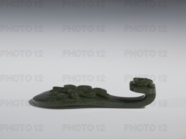 Belt clasp. Scepter head with a bas-relief stylized lizard; shaft with a Taoist emblem of one of the Eight Immortals. Spinach jade. Sculpture, engraving, 18th–19th century China. Chinese art. Chinese culture. Taoism. Qing 1644–1911. Subject: lizards; ruyi auspicious scepter; Immortals. Belt buckle. Height 2.7 cm; width 12.0 cm; depth 4.5 cm., Purchase provenance., A carved dark green object shaped with a flat rounded base and a curved raised handle, the surface decorated with carved floral motifs and rounded petal forms, shown against a smooth gradient gray background with soft shadows beneath; visible colors include dark green, lighter green highlights, gray, and white.