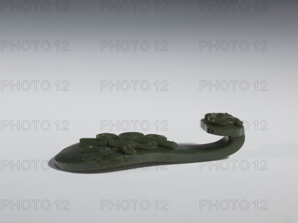 Belt clasp, Chinese, Qing 1644–1911, 18th–19th century. Spinach jade; sculpture and engraving. Scepter head with a bas-relief stylized lizard; shaft with a Taoist emblem of one of the Eight Immortals and ruyi auspicious scepter iconography; Immortals and Taoism themes; lizards zoological motif. Belt buckle. Height 2.7 cm; width 12.0 cm; depth 4.5 cm., A carved object made of dark green material rests on a light surface with a gradient gray background, showing smooth and glossy finishes and a few areas of subtle translucence; the piece has an elongated base with raised carved floral motifs and a curved extension that rises and ends in a rounded platform also decorated with carved flower shapes, casting a soft shadow beneath.