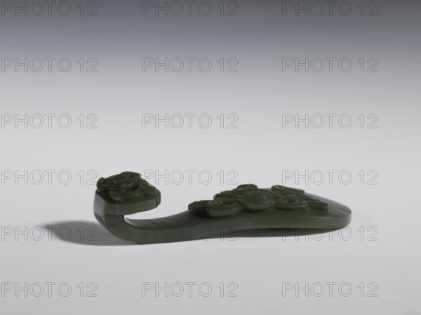 Belt clasp., Chinese. Spinach jade. Sculpture, engraving. Qing dynasty 18th–19th century. Scepter head with a bas-relief stylized lizard; shaft with a Taoist emblem of one of the Eight Immortals; ruyi auspicious scepter motif; lizards iconography; Taoism. Belt buckle. Height 2.7 cm; width 12.0 cm; depth 4.5 cm., A dark green carved object rests on a light gray surface against a gradient gray background, showing a curved form with a raised rounded end and a hooked protrusion at one side. The piece has several rounded, coiled decorative elements carved into its top surface and a smooth underside, casting a soft shadow on the surface. The visible colors are dark green, light gray, and darker gray in the background and shadow.