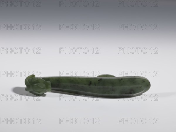 Belt clasp, Chinese, dragons iconography, Qing 1644–1911, 18th–19th century, China, spinach jade with black inclusions, slightly concave shaft surfaces, full-sculpture carved head, belt buckle, height 2.2 cm, width 10.8 cm, depth 1.7 cm, A smooth elongated object made of a green stone with darker green and black mottling rests on a light surface, one end carved into a rounded decorative form with curved, petal-like details, while the opposite end tapers into an oval shape; the background transitions from light gray near the surface to darker gray toward the top, and the object casts a soft shadow on the surface.