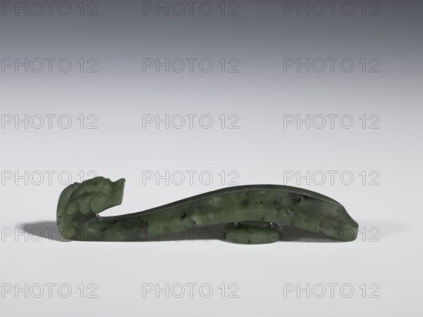 Belt clasp, Chinese, dragons iconography, Qing 1644–1911, 18th–19th century, China, spinach jade with black inclusions, slightly concave shaft surfaces, full-sculpture carved head, belt buckle; height 2.2 cm, width 10.8 cm, depth 1.7 cm, A long carved object made of green stone with darker speckles rests on a white surface against a gray gradient background; one end of the object is shaped into a stylized animal head with carved curves and rounded features, the body tapers and arches slightly and is supported near the middle by a circular stone ring, and the overall colors visible are various shades of green, dark green, white, gray, and black.