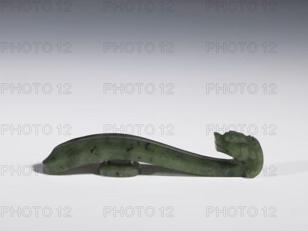 Belt clasp, Chinese, dragons iconography, Qing 1644–1911, 18th–19th century, China, spinach jade with black inclusions, slightly concave shaft surfaces, full-sculpture carved head, belt buckle, height 2.2 cm, width 10.8 cm, depth 1.7 cm., A smooth green stone object with darker green flecks rests horizontally on a light surface, supported by a small rounded base; one end tapers and curves upward into a carved form resembling an animal head with a defined snout and folded ear, while the opposite end is rounded and slightly flattened, and the overall scene shows shades of green, dark green, light gray, and white.