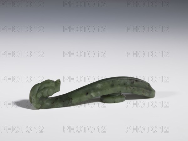 Belt clasp, Chinese, dragons iconography, Qing 1644–1911, slightly concave shaft surfaces, spinach jade with black inclusions, full-sculpture carved head, 18th–19th century, China, belt buckle; height 2.2 cm, width 10.8 cm, depth 1.7 cm, A smooth green object with darker speckles rests on a white surface, curving upward at one end into a carved, rounded shape that resembles an abstract animal head; the object has a glossy finish and sits partly on a small circular green support, casting a soft shadow, with background colors transitioning from white near the surface to gray and darker gray toward the top.