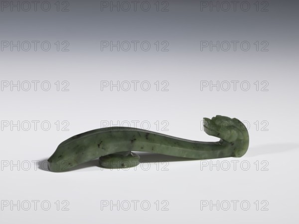 Belt clasp, Chinese, dragons iconography, Qing 1644–1911, slightly concave shaft surfaces, spinach jade with black inclusions, full-sculpture carved head, 18th–19th century, China, belt buckle, height 2.2 cm, width 10.8 cm, depth 1.7 cm, A smooth carved object made of green stone with darker flecks rests on a white surface; its shape is elongated and slightly curved with a rounded raised base beneath it, and one end rises into a stylized head-like form with subtle carved contours; colors visible include shades of green, darker green flecks, white from the surface, and gradients of gray in the background.