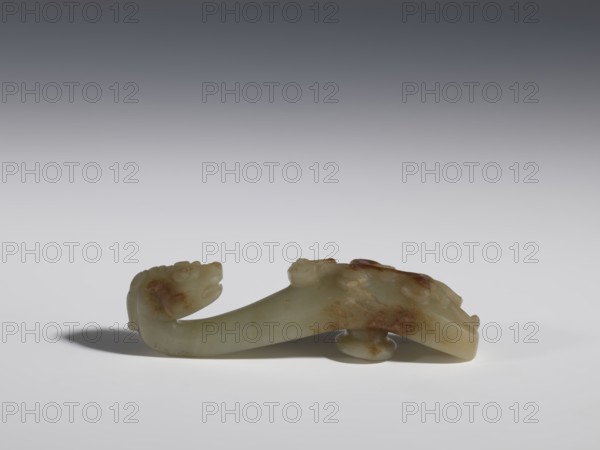 Belt clasp, Chinese, Qing 1644–1911, 17th–19th century. Jadeite with traces of chalcedony. Handle flattened on the sides, decorated with a lizard-dragon motif; head carved in full plastic, convex relief. Belt buckle. Height 2.3 cm; width 8.4 cm; depth 1.8 cm., A smooth carved object resembling a curved, elongated form with sculpted animal-like heads at each end, resting on a flat surface; its material appears jade-like with pale creamy white as the dominant color and areas of brownish and rust-colored patches; soft shadows fall beneath and to the left of the object; the background transitions from pale light gray near the surface to a darker gray toward the top.