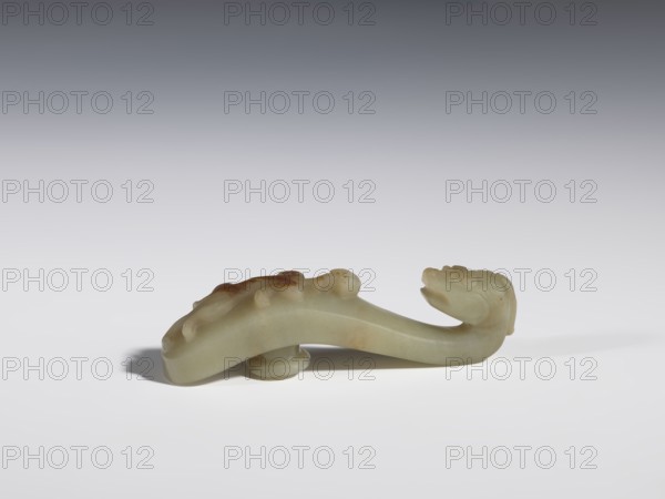 Belt clasp. Handle flattened on the sides, decorated with a lizard-dragon motif. Jadeite with traces of chalcedony. Head carved in full plastic, convex relief. Dragons; lizards. Chinese, Qing 1644–1911. 17th–19th century. Belt buckle. Height 2.3 cm; width 8.4 cm; depth 1.8 cm., A smooth sculpted object resembling a curved creature with a pronounced head at one end and a tapering body, carved from a pale greenish material with areas of darker brownish discoloration, resting on a flat white surface with a soft shadow beneath; the background fades from light gray near the surface to a darker gray toward the top, and the carving shows rounded raised details along its back and a small rounded support under its midsection.