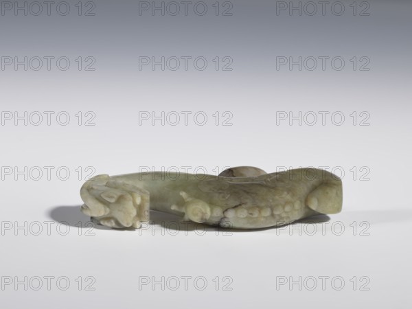 Belt clasp;, Chinese, Qing dynasty 1644–1911, 19th century. Material not specified. Head in the shape of a two-horned dragon's head with an open mouth; shaft carved as a dragon playing with a flaming pearl. Head and shaft carved in full plastic. Function: belt buckle for men's clothing/ornament. Iconography: dragons, dragon with pearl. Dimensions: height 2.65 cm; width 9.7 cm; depth 1.9 cm., A carved object rests on a flat surface, appearing to be made of a smooth, translucent material in shades of pale green, olive, and hints of brown, with variations and darker veins. The object has a curved, elongated form with rounded ends and a low, raised central area; one end features a carved motif that resembles a stylized animal head with curved, layered shapes, while the body shows a row of rounded protrusions and subtle carved lines. The surface catches soft light, creating gentle highlights and shadows, and there is a diffuse gray-to-white background behind the object.