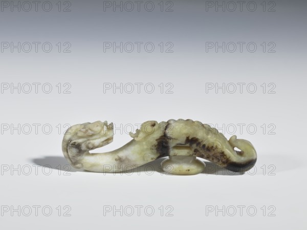 Belt clasp, Chinese, dragons iconography, Qing 1644–1911, clothing, belts, men's clothing, costumes, ornaments, men's ornaments, buckles; Head in the shape of a two-horned dragon's head with an open mouth; shaft — a dragon playing with a flaming pearl. Head and shaft carved in full plastic. 19th century, China; belt buckle; height 2.65 cm, width 9.7 cm, depth 1.9 cm., A carved object resembling a curved dragon-like creature rests on a smooth surface, showing a mix of pale green, off-white, and darker brown areas in its stone material; the piece has a rounded base beneath its midsection and detailed carved features including a curled tail and a stylized head with an open mouth, while soft shadows fall to one side against a neutral, gradient background ranging from light gray to darker gray at the top.