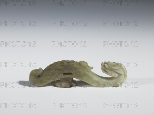 Belt clasp; maker, Chinese; dragons iconography; Qing dynasty 1644–1911; 19th century; carved in full plastic; head in the shape of a two-horned dragon's head with an open mouth; shaft depicting a dragon playing with a flaming pearl; materials for clothing/belts/men's ornaments; belt buckle; height 2.65 cm, width 9.7 cm, depth 1.9 cm., A carved object made of greenish stone with variations of pale green, olive, and cream rests on a smooth surface; the object has a sinuous, curved form with rounded ends and carved, flowing details that suggest scales or layered petals, and it casts a soft shadow beneath it on a neutral light gray background that shifts to a darker gray toward the top.