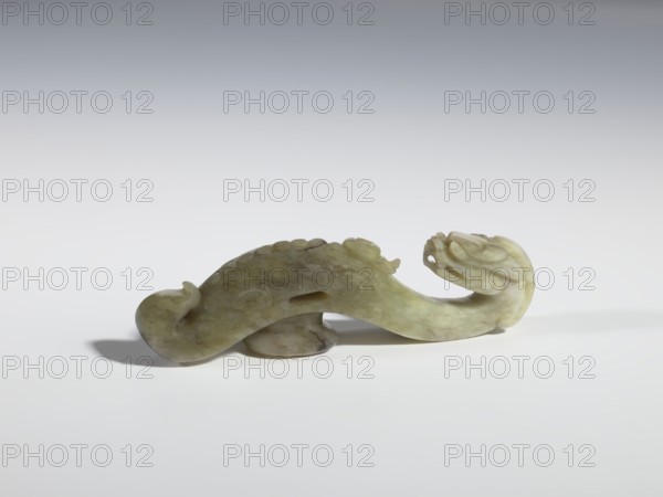 Belt clasp, purchase provenance; Chinese; Chinese art; dragons iconography; Qing 1644–1911; clothing, belts, men's clothing, costumes, ornaments, men's ornaments, buckles; Head in the shape of a two-horned dragon's head with an open mouth; shaft — a dragon playing with a flaming pearl. Head and shaft carved in full plastic. 19th century, China; belt buckle; height 2.65 cm, width 9.7 cm, depth 1.9 cm, A carved stone object in shades of pale green, light yellow, and muted brown sits on a white surface against a gradient gray background; the object has a sinuous, flowing form with a curled end resembling a stylized head with carved ridges and rounded protrusions along its back, a hollowed opening on one side, and a smooth underside that rests on a small rounded base, casting a soft shadow on the surface.