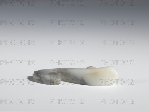 Belt clasp, Chinese, dragons iconography, Qing 1644–1911, flat profiled handle. White jadeite with gray shading, fully carved head. 17th–19th century, China. Belt buckle; height 2.1 cm, width 8.9 cm, depth 2.0 cm., A smooth, elongated carved stone object rests on a light surface with a soft shadow beneath it, showing a pale color palette of white, cream, gray, and subtle beige tones; the form tapers and curves with one end rounded and bulbous and the other end featuring carved, angular details and faceted edges, and the stone surface displays faint veins and patches of slightly darker gray and warm beige.