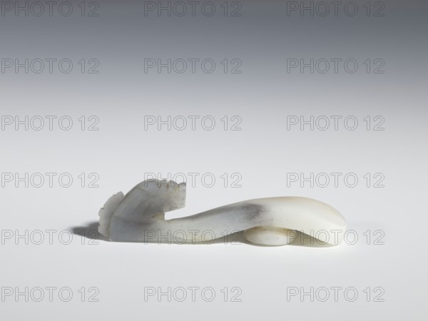 Belt clasp, Chinese, dragons iconography, Qing 1644–1911, flat profiled handle. White jadeite with gray shading, fully carved head. 17th–19th century, China. Belt buckle; H 2.1 cm, W 8.9 cm, D 2.0 cm., A smooth carved object rests on a flat surface, showing an elongated curving form with a rounded end and a carved head-like shape at the other end with ridged details; the object appears to be made of a translucent stone with areas of white, pale gray, and subtle beige, and it casts a soft shadow on the surface beneath it.