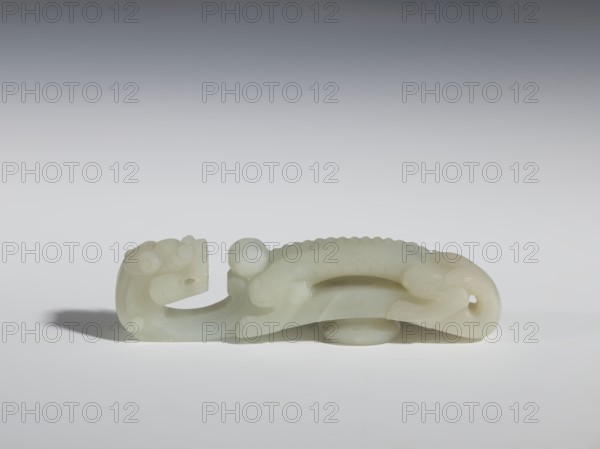 Belt clasp, Chinese, dragons iconography, dragon with pearl, Qing 1644–1911. Head in the shape of a two-horned dragon's head with an open mouth; stem - a dragon playing with a flaming pearl. Dark milky jadeite, translucent with inclusions dirty green. Head and stem carved in full relief. 19th century, China. Belt buckle; height 3.1 cm, width 11.6 cm, depth 2.3 cm., A pale, translucent carved object resting on a smooth surface, shaped like a stylized animal with a rounded head, curled tail, and a sinuous body; the carving shows scalloped ridges along the back, rounded protrusions near the head, and flowing, curved lines suggesting limbs and facial features. The object is primarily milky white with subtle variations of cream and very light greenish tones, and it casts a soft gray shadow on the surface beneath it. The background transitions from light gray at the bottom to darker gray toward the top.