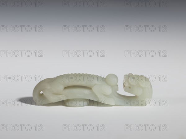 Belt clasp, Chinese, dragons iconography, dragon with pearl, Qing 1644–1911. Head in the shape of a two-horned dragon's head with an open mouth; stem - a dragon playing with a flaming pearl. Dark milky jadeite, translucent with inclusions dirty green. Head and stem carved in full relief. 19th century, China. Belt buckle. Height 3.1 cm; width 11.6 cm; depth 2.3 cm., A pale, smooth carved object resembling an elongated creature rests on a light surface; the carving is off-white to creamy with subtle translucent areas and a slightly glossy finish, featuring rounded forms including a bulbous head, curved body with scalloped ridges along its back, and a curled tail with a hole near its tip; the piece casts a soft gray shadow beneath and to one side, and the background transitions from pale gray near the surface to darker gray toward the top.