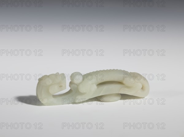 Belt clasp, Chinese, dragons iconography, dragon with pearl, Qing 1644–1911. Head in the shape of a two-horned dragon's head with an open mouth; stem a dragon playing with a flaming pearl. Dark milky jadeite, translucent with inclusions dirty green. Head and stem carved in full relief. 19th century, China. Belt buckle. Height 3.1 cm; width 11.6 cm; depth 2.3 cm., A pale, smooth carved object resembling a stylized animal form rests on a flat surface, showing a curved body with a rounded head at one end and a tapered tail at the other; the carving has sinuous contours, a raised ridge along the back with scalloped detailing, rounded protrusions and curving open spaces within the body, and a separate rounded piece beneath it providing slight elevation; the visible colors are shades of off-white, pale cream, and very light gray, set against a background that transitions from light gray at the bottom to darker gray toward the top.