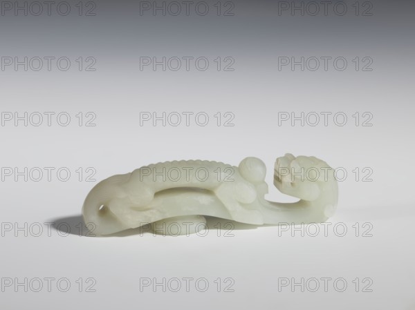Belt clasp, Chinese, dragons iconography, dragon with pearl, Qing 1644–1911. Head in the shape of a two-horned dragon's head with an open mouth; stem a dragon playing with a flaming pearl. Dark milky jadeite, translucent with inclusions dirty green. Head and stem carved in full relief. 19th century, China. Belt buckle. Height 3.1 cm, width 11.6 cm, depth 2.3 cm., A pale greenish-white carved stone object in the shape of a stylized animal with a long curved body and a carved head rests on a light surface, casting a soft shadow; the object shows smooth rounded forms, textured ridges along its back, a curled tail with a small hole, and subtle variations of creamy, off-white, and translucent green tones.